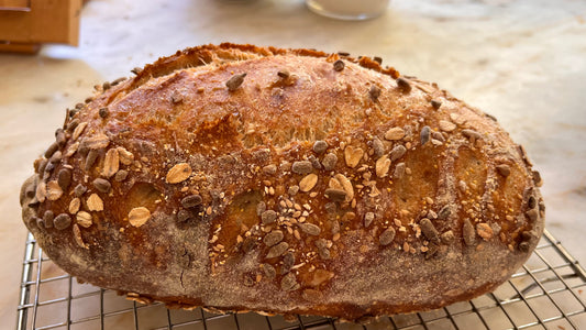 It Begins with Bread: A golden seed-crusted sourdough loaf cooling on a wire rack — handmade bread baked from scratch in a Santa Barbara hands-on cooking class